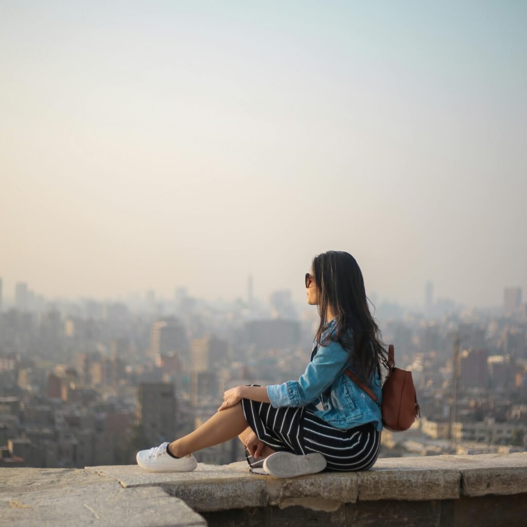 A young woman in casual attire sits on a rooftop terrace, viewing a sprawling cityscape in daylight.