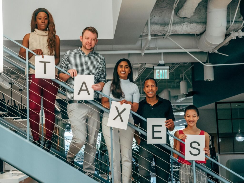 pexels-photo-7688525-7688525 A diverse group of professionals smiling and holding 'TAXES' signs on an indoor stairway
